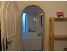 Geöffnete, weiße Tür mit bogenförmigem Rahmen führt in ein kleines Badezimmer. Das Bad hat weiße Fliesen, eine Badewanne mit Duschwand und ein Fenster mit milchigem Glas; daneben steht ein offenes Holzregal mit dekorativen Objekten.