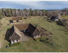 Luftaufnahme eines historischen Gehöfts mit Fachwerkhäusern und Reetdächern in ländlicher Umgebung. Die Gebäude sind von offenen Wiesen, einzelnen Bäumen und einem angrenzenden Wald umgeben.