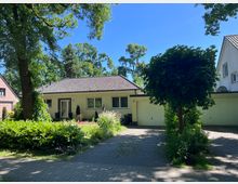 Einfamilienhaus mit flachem Walmdach und heller Fassade in einer grünen Wohnstraße, davor Gartenbeete und ein gepflasterter Zufahrtsweg. Rechts schließt eine Garage an, umgeben von großen Bäumen und dichtem Strauchwerk.