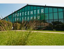 Großes Gebäude mit Glasfassade, beschriftet mit "Sportcenter Wittenau", umgeben von einer gepflegten Rasenfläche und hohen Gräsern im Vordergrund. Im Hintergrund sind Büsche und Bäume unter klarem Himmel sichtbar.