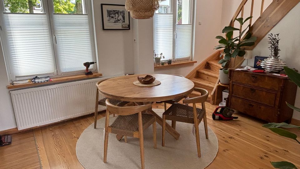 Heller Essbereich mit rundem Holztisch und vier Stühlen auf einem runden Teppich, daneben eine Holztreppe mit Geländer. Große Fenster mit halb geschlossenen Plissees, eine geflochtene Hängelampe sowie eine Kommode mit Pflanzen stehen an den Wänden.