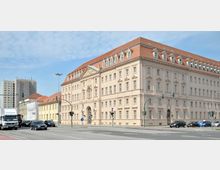 Historisches Gebäude mit heller Fassade und rotem Satteldach, das von einem symmetrischen Stil mit vielen Fenstern und einer großen Eingangstür geprägt ist, an einer mehrspurigen Kreuzung mit Ampeln und vorbeifahrenden Autos gelegen. Im Hintergrund sind ältere und moderne Gebäude zu sehen.