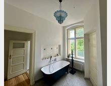 Helles Badezimmer mit einer freistehenden Badewanne und Wandarmatur vor einer gefliesten Nische. Rechts ein großes Fenster mit Blick ins Grüne, darunter ein Heizkörper; links ein offener Durchgang mit einer weißen Tür.