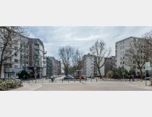 Straßenszene in einer Wohngegend mit modernen und älteren Wohngebäuden, flankiert von Bäumen ohne Laub. Im Vordergrund ein öffentlicher Platz mit Pollern, geparkten Fahrrädern und einem Mülleimer, im Hintergrund parkende Autos und Fußgänger.