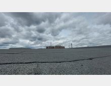 Flaches Dach mit einer Kies- oder Bitumenoberfläche, im Hintergrund ein großes Backsteingebäude mit Fenstern und zwei Mobilfunkantennen, darüber ein bewölkter Himmel.