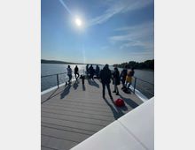 Eine Gruppe von Menschen steht auf dem Deck eines Bootes, mit Blick auf ein ruhiges Gewässer und bewaldete Ufer im Hintergrund. Die Sonne scheint hell am blauen Himmel, und lange Schatten der Personen fallen auf den Boden des Decks.