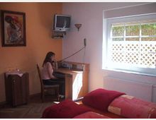 Ein kleines Schlafzimmer mit einem Schreibtisch an der Wand, auf dem ein Telefon und ein alter Röhrenfernseher stehen. Neben dem Schreibtisch sitzt eine Frau, während ein Bett mit roten Decken im Vordergrund zu sehen ist und Tageslicht durch ein großes Fenster mit Gitterstruktur einfällt.