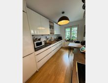 Moderne Küche mit glatten, weißen Einbauschränken und einer dunklen Arbeitsplatte, auf der verschiedene Küchengeräte stehen. Der Raum hat einen Holzfußboden, ein großes Fenster mit Pflanzen auf der Fensterbank und eine zentrale Hängeleuchte mit rundem Schirm.