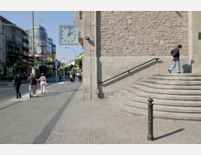 Straßenszene in einer städtischen Umgebung mit einer Treppe aus Stein, die zu einem Gebäude mit heller Steinfassade führt. Eine große Uhr ist an der Gebäudeaußenwand befestigt, dahinter sind Geschäfte und das Einkaufszentrum "Neukölln Arcaden" zu sehen.