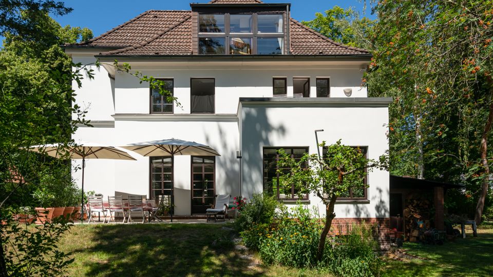 Rückansicht eines zweigeschossigen weißen Wohnhauses mit braunem Ziegeldach und großer Gaube mit Fensterfront, umgeben von Bäumen. Im Garten davor liegen eine Terrasse mit Sonnenschirmen und Sitzmöbeln sowie Rasen, Sträucher und ein kleiner Baum.
