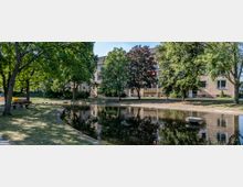 Wohnanlage mit gepflegtem Park und Teich. Im Vordergrund steht ein kleiner See mit Bäumen und Sitzbänken entlang des Ufers, im Hintergrund befinden sich beige Wohngebäude mit Balkonen.