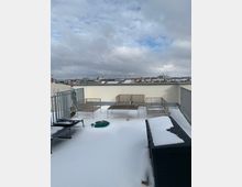 Schneebedeckte Dachterrasse mit minimalistischen Metallmöbeln, darunter ein Tisch und Sitzbänke, sowie blick auf eine Stadtlandschaft mit Wolken und vereinzeltem blauem Himmel im Hintergrund.