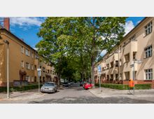 Straßenszene in einem Wohngebiet mit zweigeschossigen, gelben Wohnhäusern im Stil der 1920er bis 1930er Jahre, roten Backsteinfassaden und zahlreichen Fenstern. Bäume säumen die Straße, auf der Autos geparkt sind, und es sind Straßenschilder sowie ein orangefarbener Briefkasten zu sehen.