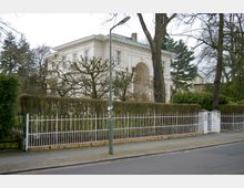 Weiße zweigeschossige Villa mit klassizistischen Details und einem großen Rundbogenportal, umgeben von einer Hecke und einem weißen Metallzaun, vor einer Baumreihe; im Vordergrund eine Straße mit Gehweg und Straßenlaterne.