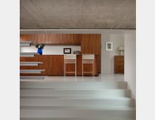 Interior view of a modern home bar area with floor-to-ceiling wood cabinetry, a long counter, and two white bar stools on a light terrazzo floor. In the foreground are wide white steps and a floating staircase with gray treads, set beneath an exposed concrete ceiling.