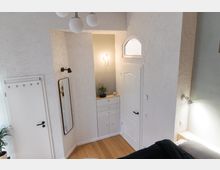 Heller Innenraum eines Zimmers mit weiß verputzten Wänden, einem eingebauten Schrank mit Holzplatte und einer beleuchteten Nische. Ein weißes Bogentürfenster, eine schmale Zimmertür, ein rechteckiger Spiegel und moderne Leuchten ergänzen das schlichte Design.