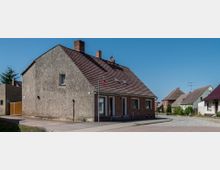 Straßenszene in einer ländlichen Siedlung mit einem niedrigen Wohnhaus aus verputztem Mauerwerk und rotem Ziegeldach im Vordergrund. Im Hintergrund stehen weitere ähnliche Häuser, teils mit Dachfenstern, sowie kleinere Gehwege und eine gepflasterte Straße.