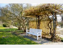 Weiße Parkbank unter einer metallenen Pergola mit Kletterpflanzen, daneben ein Baum und umliegende Grünflächen; im Hintergrund eine Gehwegkreuzung in einem Garten.