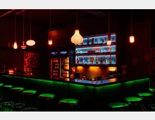Dunkle Bar mit rechteckiger Theke, beleuchtet durch grüne LED-Lichter an der Bar und warmen Hängelampen in verschiedenen Formen. Im Hintergrund befinden sich beleuchtete Getränkeregale mit Flaschen sowie zwei Getränkekühlschränke.