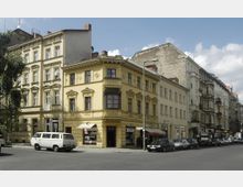 Gelbe und beigefarbene Wohngebäude aus der Gründerzeit an einer Straßenecke in einer Stadt, mit Fenstergesimsen und dezenten Stuckverzierungen. Mehrere parkende Autos und ein Lieferwagen befinden sich entlang der Straße, die von einem Baum und einer Straßenlaterne gesäumt wird.