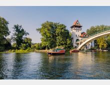 Ansicht eines Flusses mit Booten, einer weißen Bogenbrücke rechts und einem historischen Gebäude mit einem roten Spitzdach im Hintergrund, umgeben von Bäumen. Auf dem Gebäude steht "Insel Berlin".