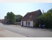 Eine Dorfstraße mit einem kleinen, älteren Gebäude auf der rechten Seite, das teils verputzt und teils aus Holz besteht, mit einem Fahrradgeschäftsschild. Daneben stehen Fahrräder, ein Motorroller und Bäume, die das Gelände umgeben.