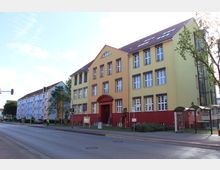 Gelb-rotes Gebäude einer Musik- und Kunstschule mit großen Fenstern und einer Eingangstür im Bogendesign, daneben eine überdachte Bushaltestelle. Im Hintergrund sind mehrstöckige, pastellfarbene Wohnhäuser zu sehen, die an einer Straße mit Gehweg liegen.