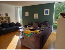 Helles Wohnzimmer mit Holzboden, einem großen, L-förmigen grauen Sofa und einer dunkelgrünen Wand mit drei Bildern. Links steht ein Bücherregal vor einer weißen Wand, rechts sind bodentiefe Fenster mit Blick auf Bäume.