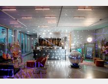 Moderner Veranstaltungsraum mit einer Bar im Hintergrund, dekoriert mit Pflanzen und Blumen. Im Vordergrund stehen Sitzmöbel, ein glitzernder Discoball und ein leuchtender Spiegel, während bunte Lichtpunkte von der Decke projiziert werden.