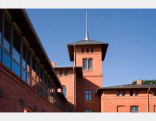 Ziegelrotes Gebäude mit einem zentralen Turm und spitzem Dachaufsatz, flankiert von weiteren Gebäudeteilen mit großen Fenstern; der Himmel ist klar und blau, einige Vögel sind sichtbar.
