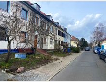 Reihenhäuser in einer ruhigen Wohnstraße mit gepflastertem Gehweg, Vorgärten und Bäumen im kahlen Winterzustand. Geparkte Autos säumen die Straße, und blauer Himmel mit Wolken ist im Hintergrund zu sehen.