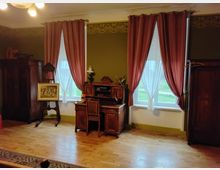 Ein Zimmer mit Vintage-Möbeln, darunter ein Schreibpult, zwei große Holzschränke und ein Ständer mit einem gerahmten Bild. Zwei Fenster mit rosa Vorhängen und weißen Gardinen lassen Tageslicht hinein; der Raum hat einen holzfarbenen Boden und eine grün gestrichene Wand mit dekorativer Bordüre.