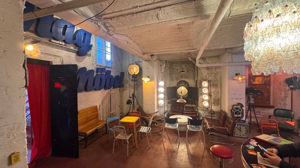 Gemütlicher Raum mit industriellem Look, weiß gestrichenen Backsteinwänden und sichtbaren Rohren an der Decke. Der Raum ist mit einer Mischung aus Vintage-Möbeln, bunten Stühlen, roten Barhockern und dekorativen Lichtelementen eingerichtet, einschließlich eines Kronleuchters und alter Filmbeleuchtung.