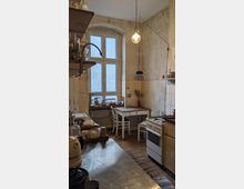 Rustikale Küche mit einem hohen Bogenfenster, Holzboden und abgenutzten, verputzten Wänden. Im Raum befinden sich ein Herd, eine Arbeitsfläche aus Edelstahl, ein kleiner Esstisch mit zwei Stühlen sowie hängende Lampen, die warmes Licht spenden.