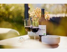 Eine Weinflasche mit der Aufschrift "Mouton Cadet Vin Bio 2021" steht auf einem Tisch zusammen mit zwei gefüllten Weingläsern, einer Schale und einem Korkenzieher, im Hintergrund unscharfe Pflanzen und Lichtreflexionen.