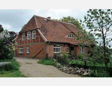 Backsteinhaus mit rotem Ziegeldach, einem geschwungenen Giebel und mehreren weißen Fenstern. Das Haus steht in einer grünen Umgebung mit Bäumen, Sträuchern und einem kleinen Garten, entlang eines unbefestigten Weges.