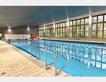 Innenansicht eines Hallenbades mit einem großen Schwimmbecken, das von Startblöcken und Edelstahltreppen umgeben ist. Die hohen Fenster auf der rechten Seite lassen Tageslicht herein und bieten Blick auf Bäume im Außenbereich.