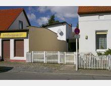Straßenszene mit zwei Gebäuden: Links ein Laden mit der Aufschrift „Bastlerbedarf“, geschlossenen Rollläden und rotem Ziegeldach; rechts ein Wohnhaus mit weißer Fassade, einem Fenster und einem kleinen Vorgarten mit weißem Holzzaun. Zwischen den Gebäuden befindet sich ein schmaler, eingefasster Durchgang.