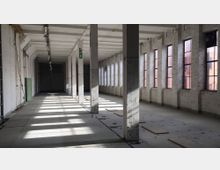 Leerstehender industrieller Innenraum mit weißen Backsteinwänden, rechteckigen Fenstern und einer Reihe von Säulen, die den Raum in zwei Abschnitte teilen. Sonnenlicht fällt durch die Fenster, wodurch Schatten auf den Boden geworfen werden.