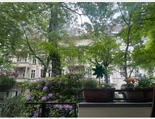 Blick von einem Balkon auf einen begrünten Innenhof mit blühenden Blumen in Töpfen im Vordergrund. Im Hintergrund sind weiße Wohngebäude mit Balkonen und großen Fensterfronten, umgeben von hohen, dicht belaubten Bäumen, sichtbar.