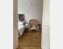 Helles Schlafzimmer mit Fischgrät-Holzboden, einem Bett mit weißer Bettwäsche und einem dekorativen Korbsessel mit Fellauflage. Im Raum stehen außerdem ein kleiner Beistelltisch mit Tischlampe, eine Zimmerpflanze und ein Gemälde an der Wand.