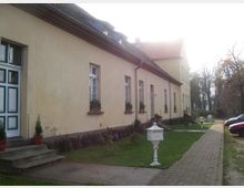 Ein langes, gelbes Gebäude mit mehreren weißen Fenstern, teilweise mit Blumenkästen versehen. Vor dem Gebäude befinden sich eine gepflasterte Gehwegfläche, Rasenflächen mit kleinen Dekolaternen sowie einige geparkte Autos am rechten Bildrand.