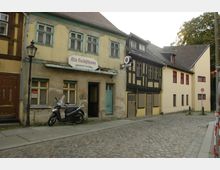 Eine kopfsteingepflasterte Straße mit historischen Gebäuden, darunter ein zweistöckiges, gelblich-beiges Haus mit der Aufschrift "Alte Volksschenke – erbaut um 1231". Davor steht ein Motorroller und eine traditionelle Laterne; daneben ein Fachwerkhaus mit hellen Mauern und dunklen Holzbalken.