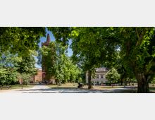 Historischer Park mit einem mittelalterlichen Backsteinturm im Zentrum, umgeben von großen Bäumen. Im Hintergrund ist ein helles Gebäude im klassischen Stil zu sehen, und Wege führen durch die grüne Anlage.
