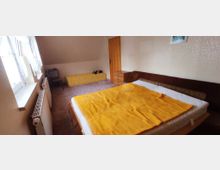 Schlafzimmer mit einem Einzelbett mit gelber Decke, einer braunen Couch, einem kleinen Fernseher auf einem Tisch, einem gelben Sideboard und Heizkörpern unter einem Fenster mit Spitzenvorhängen. Wände sind beige, der Boden ist mit Teppich ausgelegt.