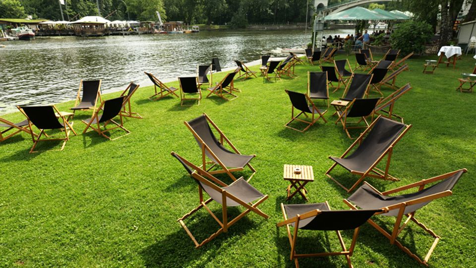 Rasenfläche am Ufer eines Flusses mit zahlreichen Liegestühlen und kleinen Beistelltischen, teils unter einem Sonnenschirm. Im Hintergrund sind Bäume sowie eine Uferpromenade mit weiteren Sitzbereichen zu sehen.