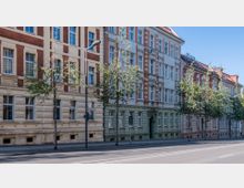 Straßenansicht mit einer Reihe von mehrstöckigen Wohngebäuden im klassischen Baustil mit Verzierungen und Klinkerfassaden; Bäume säumen den Gehweg entlang der ruhigen Straße, die mit Laternen ausgestattet ist.