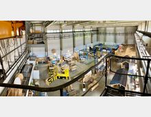 Große Werkhalle mit hohen Decken und Metallträgern, gefüllt mit Arbeitsstationen, Regalen, Materialien und verschiedenen Bauprojekten. Die Halle ist hell beleuchtet durch große Fenster an den Wänden.