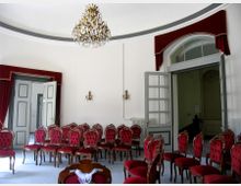 Ein eleganter, heller Raum mit einer gewölbten Decke, verziert mit einem Kronleuchter. Der Raum ist mit roten, gepolsterten Stühlen mit Holzrahmen eingerichtet, und große Doppeltüren, umrahmt von roten Vorhängen, führen zu anderen Bereichen.