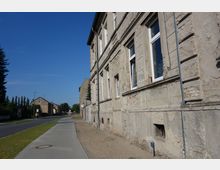 Eine alte, teils verfallene Hausfassade mit sichtbarem Putz- und Mauerwerksschäden steht an einer ruhigen Straße mit Bürgersteig. Im Hintergrund sind weitere Häuser, Bäume und ein blauer Himmel sichtbar.
